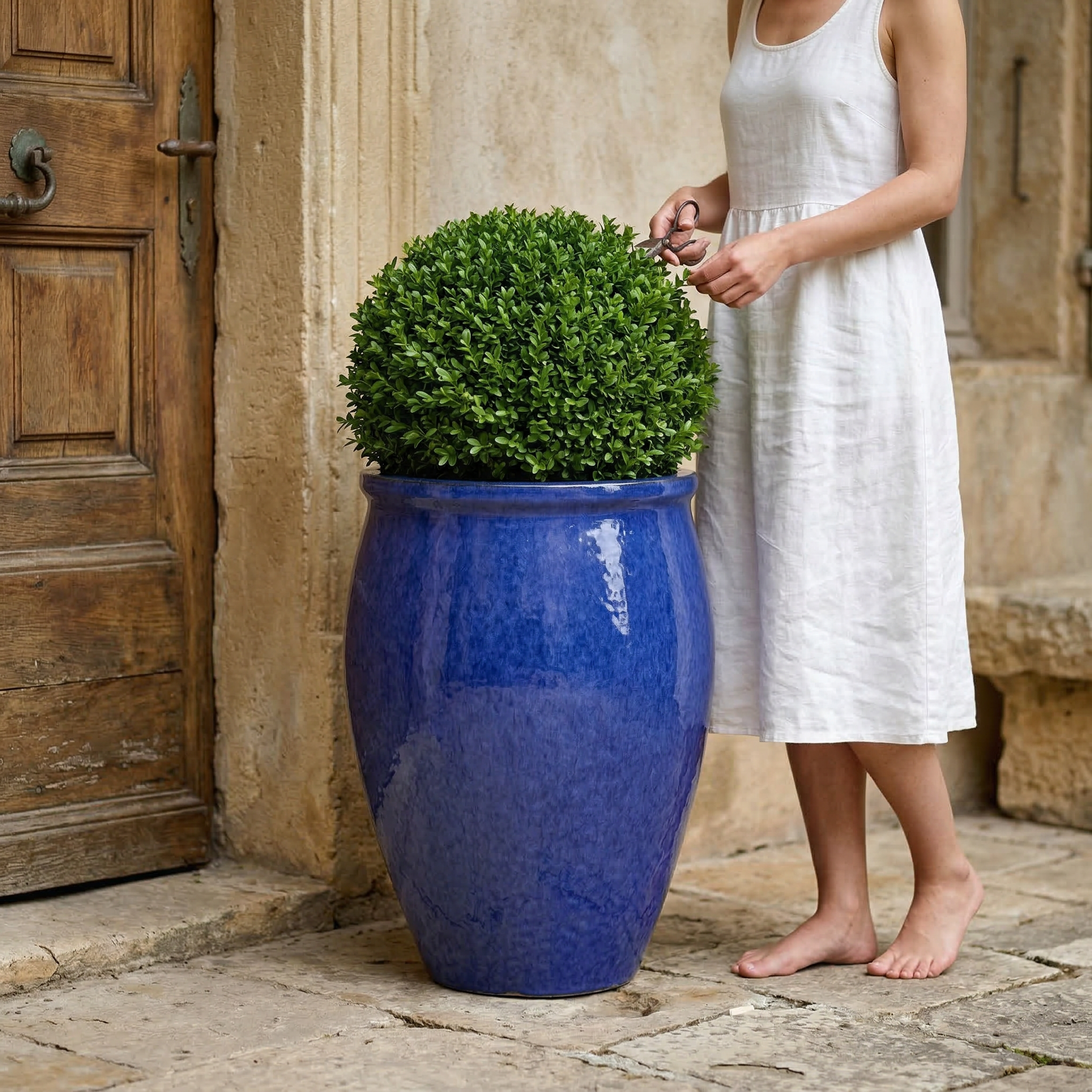 Keramický květináč BACCHUS - Majorellová modř s kulatým buxusem před starými dřevěnými dveřmi. Žena v letních bílých šatech pečuje o rostlinu.
