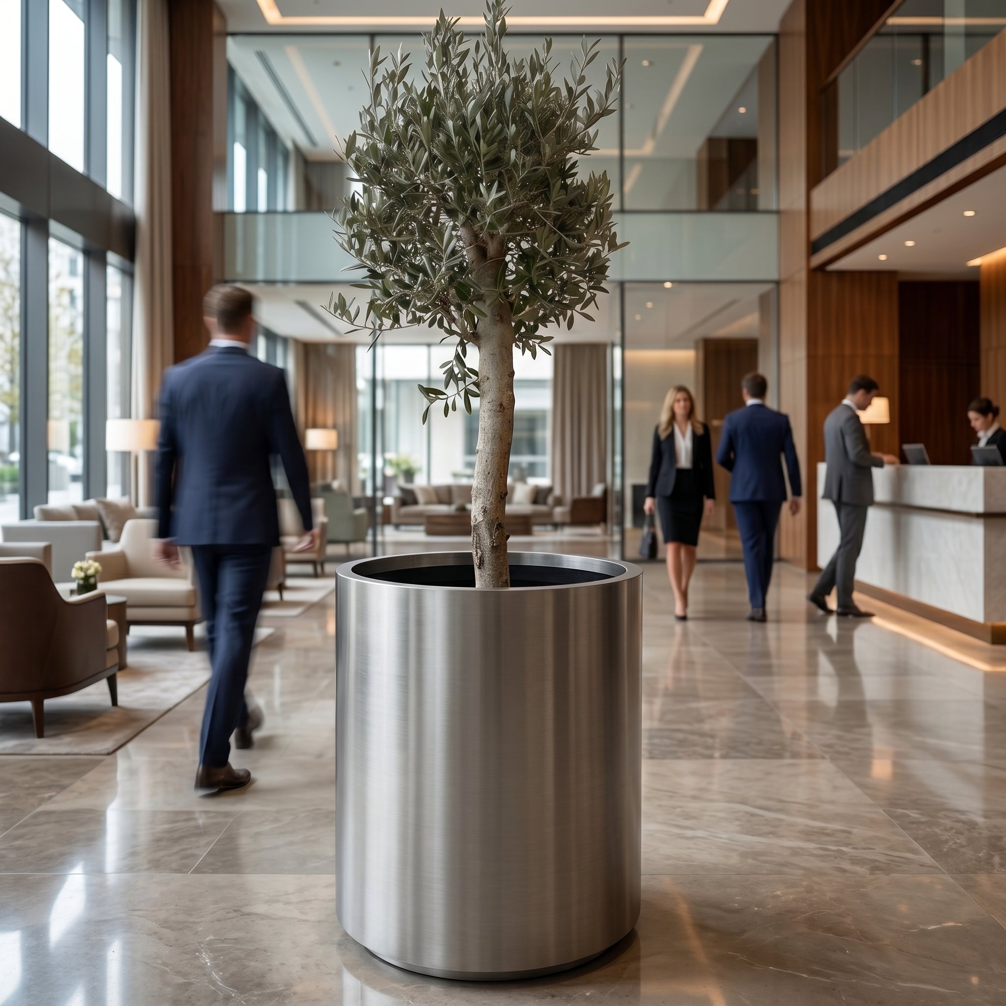 Olivovník (silný kmen) v černém designovém květináči METROPOLE 60×70 cm jako dominanta luxusního hotelového lobby s procházejícími hosty.